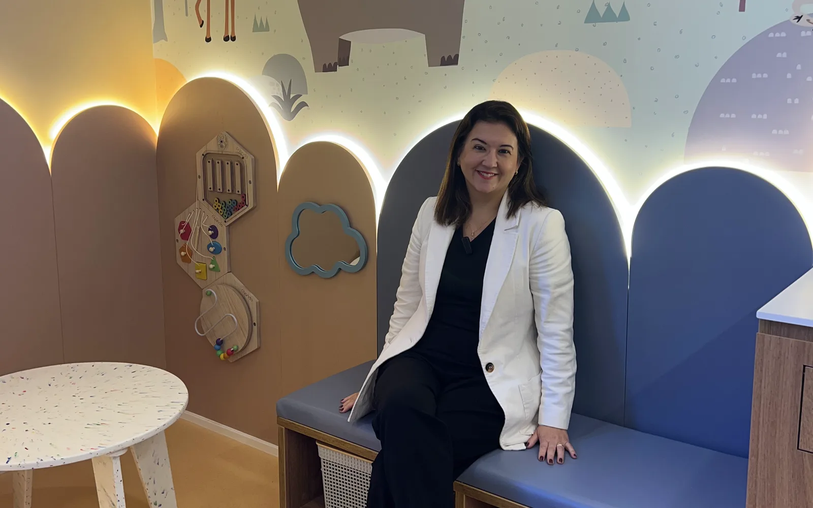 Dra. Adriane Yamamoto sentada no consultório do Ibirapuera Kids, diante de um painel com desenhos de animais e brinquedos de parede
